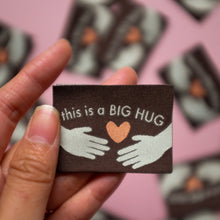 Load image into Gallery viewer, Close-up of a hand holding a brown woven label with the text "This is a BIG HUG" in white, featuring two illustrated hands reaching toward an orange heart in the center. The blurred background includes additional woven labels scattered on a pink surface.