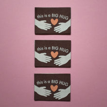 Load image into Gallery viewer, 3 identical woven labels laid flat on a pink background. Each label is brown with the text "This is a BIG HUG" in white, featuring two illustrated hands reaching toward an orange heart in the center.