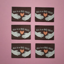 Load image into Gallery viewer, Six identical woven labels laid flat on a pink background. Each label is brown with the text "This is a BIG HUG" in white, featuring two illustrated hands reaching toward an orange heart in the center.