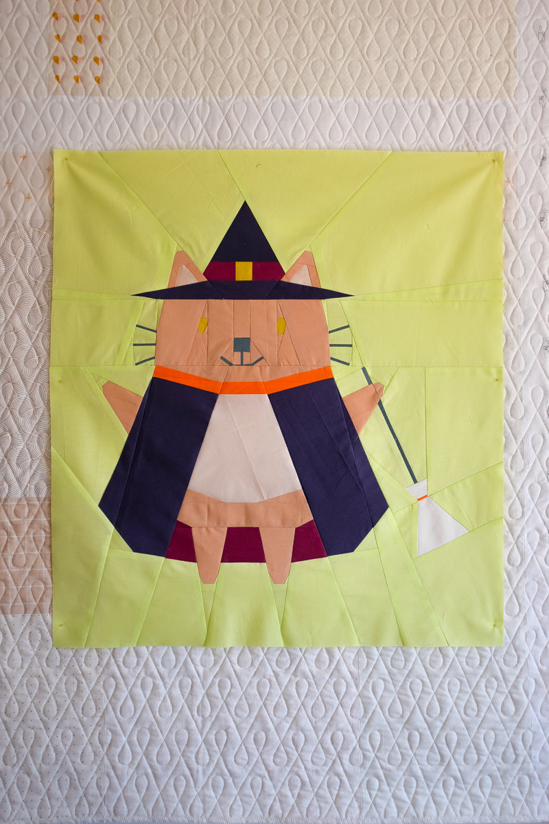 Quilt block with a cat in a witch costume on a textured wall. The Weekend Quilter Halloween Catwalk Series Hattie Cat Mini Quilt Pattern