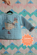 Load image into Gallery viewer, A light blue quilted jacket with a duck patchwork design hanging on a black hanger, held by a hand against a colorful quilted background. An orange starburst overlay reads 'Inspiration: Quilt block on quilt jacket'. The Weekend Quilter Frosty Flappers quilt block (Beaks on Ice Series)