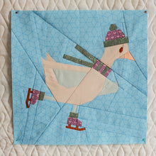 Load image into Gallery viewer, A square quilted fabric block featuring a stylized duck wearing a patterned knit hat, scarf, and ice skates, set against a light blue geometric background, displayed on a white quilted surface. The Weekend Quilter Frosty Flappers quilt block (Beaks on Ice Series)