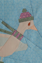 Load image into Gallery viewer, A close-up of a quilted fabric design featuring a stylized bird or duck in pale peach and muted green, wearing a pink and purple patterned hat and matching scarf, set against a light blue geometric quilted background. The Weekend Quilter Frosty Flappers quilt block (Beaks on Ice Series)