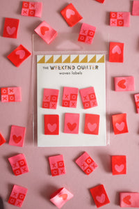 A vibrant flat lay of The Weekend Quilter's Hugs and Kisses woven labels in shades of pink, red, and pastel. The design includes "XOXO" motifs and reversible heart labels with light and dark sides, perfect for enhancing quilts, bags, and handmade sewing projects with a colorful, personalized touch.