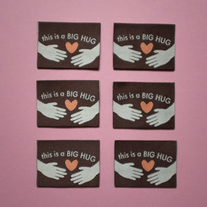 Six identical woven labels laid flat on a pink background. Each label is brown with the text "This is a BIG HUG" in white, featuring two illustrated hands reaching toward an orange heart in the center.