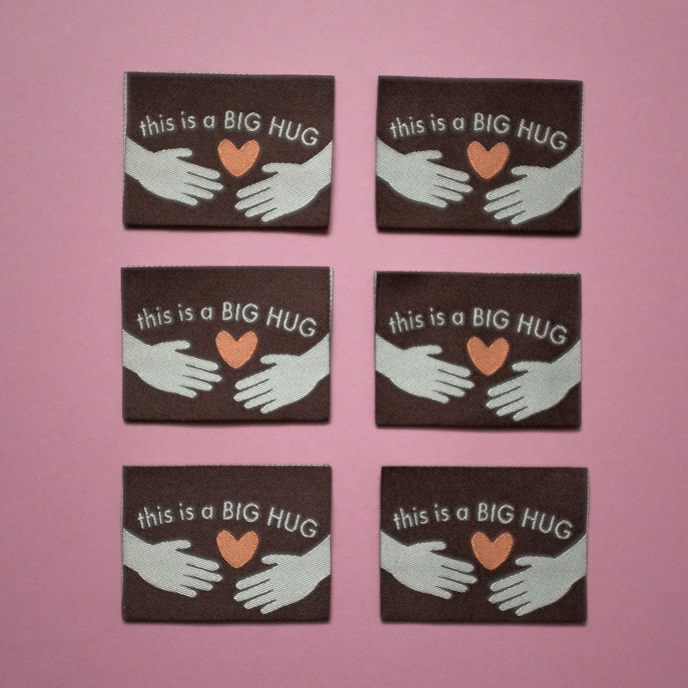Six identical woven labels laid flat on a pink background. Each label is brown with the text 