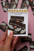 Load image into Gallery viewer, A packaged set of brown woven labels titled &quot;The Weekend Quilter Woven Labels.&quot; The label design includes the text &quot;This is a BIG HUG&quot; in white, two illustrated hands, and an orange heart. The packaging is held up by a hand against a blurred background of similar labels.